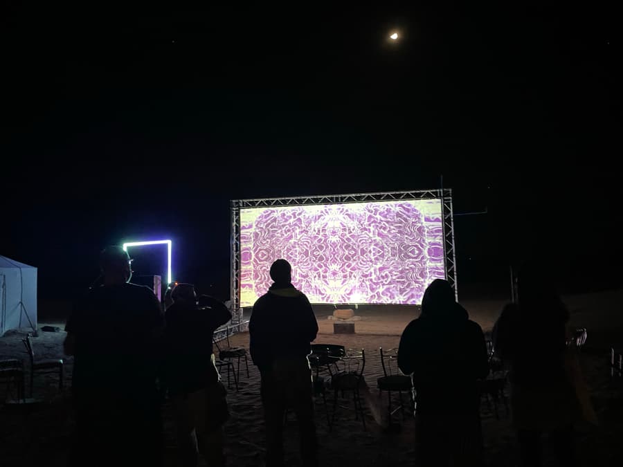 The projection screen under a full moon in the desert, with a lit frame installation alongside